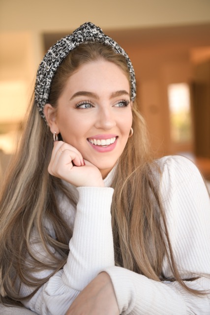 Tehilla smiling, wearing a black-and-white tweed headband and white ribbed top, resting her chin on her hand